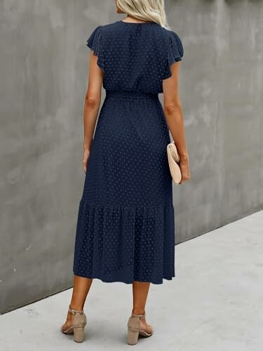 Woman in a navy blue dress with ruffled sleeves, standing against a gray wall.